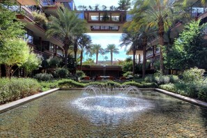 Surrounded by lush greenery and palm trees, this serene courtyard features a central water feature and multi-story buildings with balconies.