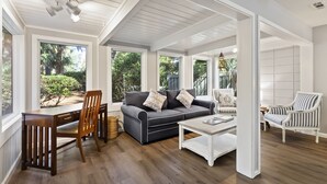 A spacious living room with a dark grey sofa, a white coffee table, and striped armchairs. There is a wooden dining table with chairs, and large windows offering views of greenery outside.