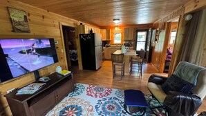 Un intérieur de chalet chaleureux comprenant un salon, une cuisine et un coin repas, avec des murs et des planchers en bois, un téléviseur à écran plat et un tapis à motifs.