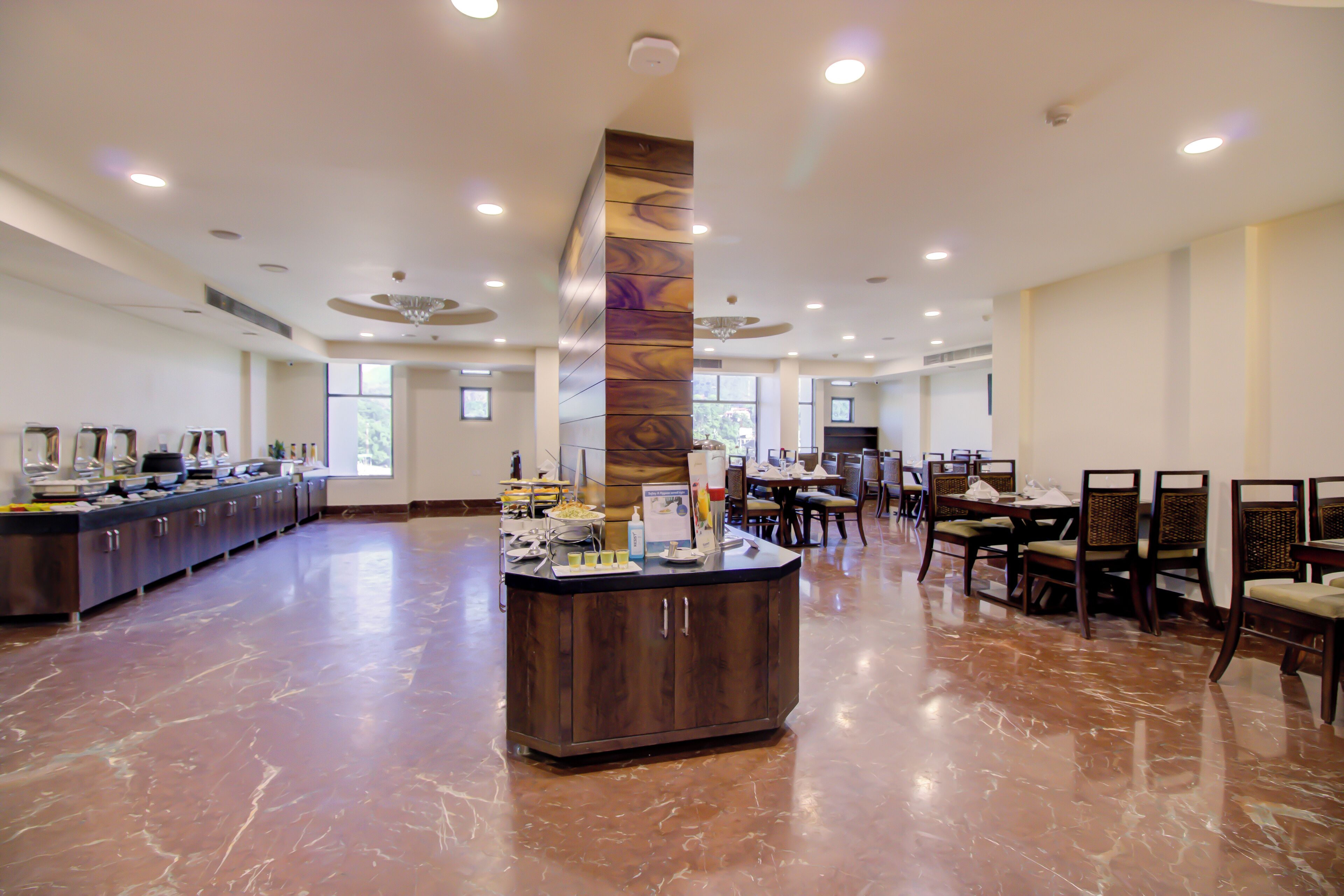 A spacious hotel lobby with a central buffet area, multiple dining tables, and chairs.