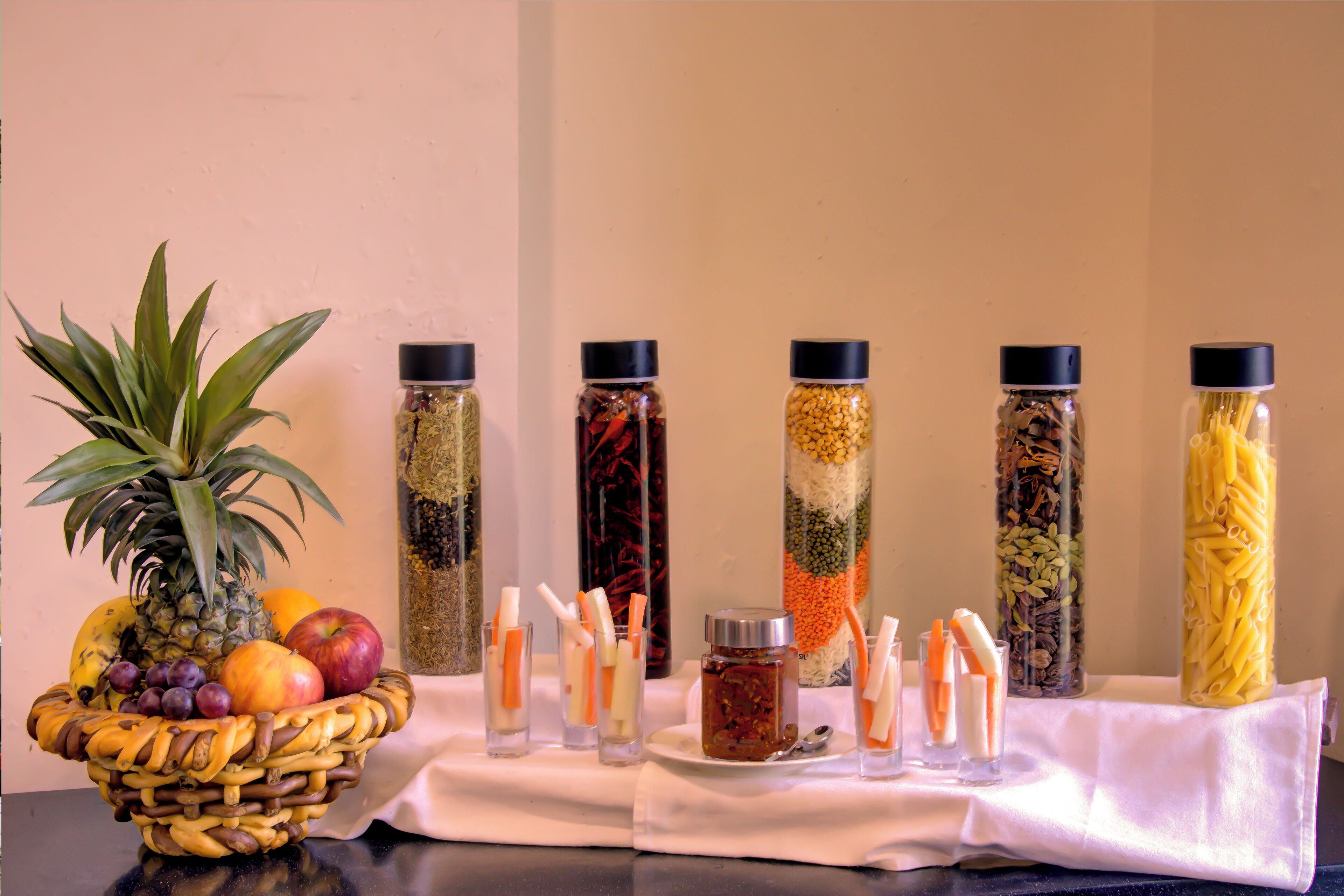 A table with a fruit basket, six spice jars, and a white cloth.