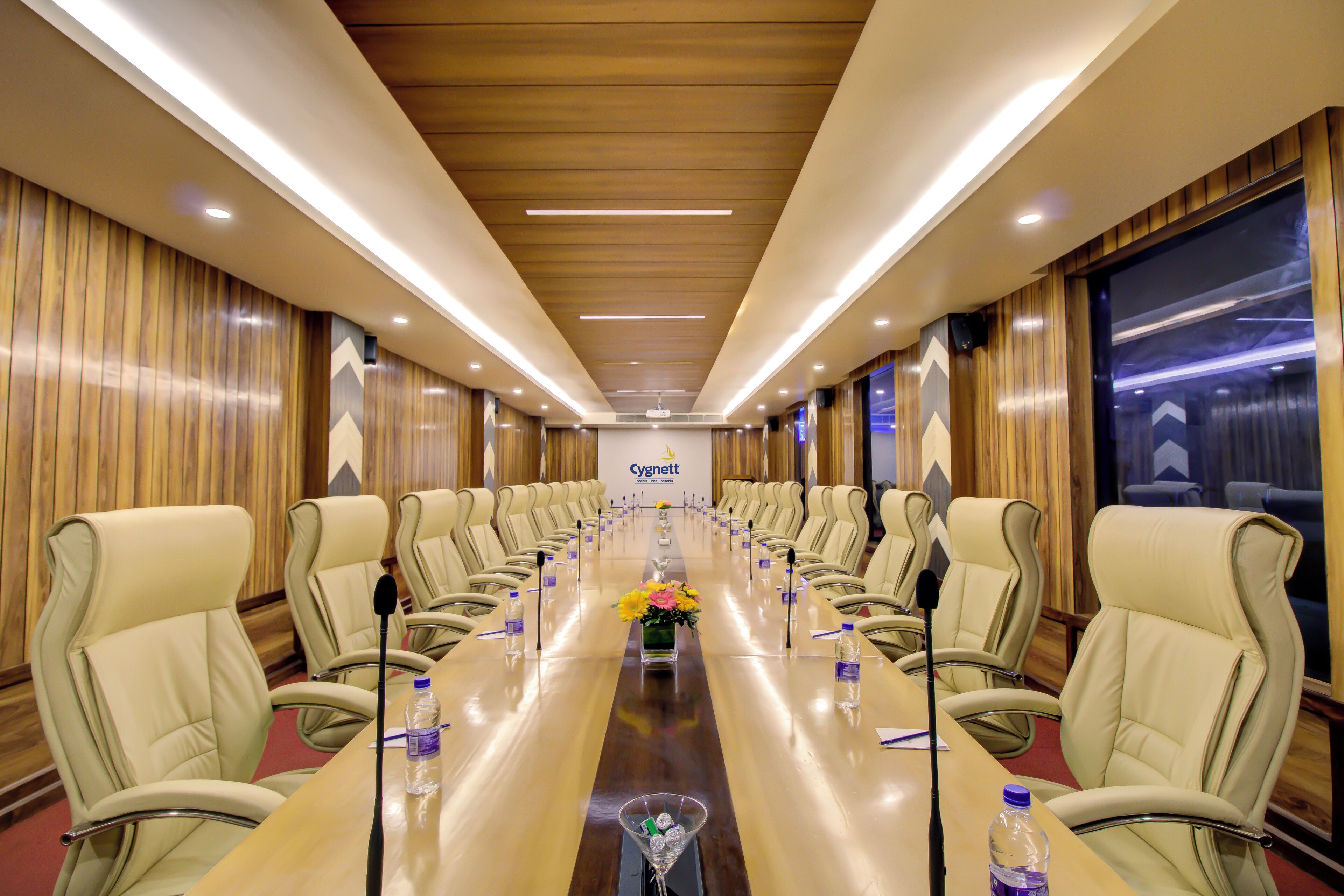 A conference room with a long table, beige chairs, microphones, water bottles, and a logo on the wall.