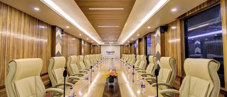 A conference room with a long table, beige chairs, microphones, water bottles, and a logo on the wall.
