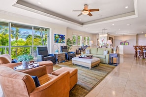 A spacious living room with leather sofas, a coffee table, and a ceiling fan. Large windows offer a view of lush greenery outside. - A 5-Star Estate, Large Groups, Luxury Living (Kihei)