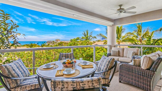 Un balcón con una mesa redonda lista para comer, rodeado de muebles de mimbre y vistas a un paisaje tropical con palmeras y el océano.