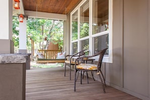A covered porch with wicker chairs and a bench, large windows, and a wooden ceiling.