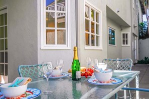 An outdoor dining area with a table set for two, a bottle of champagne, and a bowl of fruit.