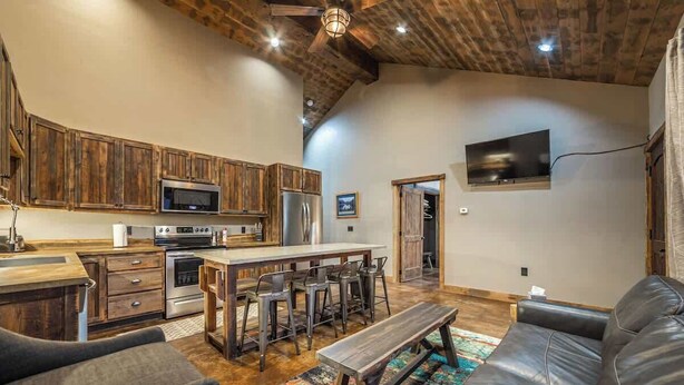 A spacious living area with a large leather sofa, a wooden dining table with bar stools, and a kitchen with wooden cabinets and modern appliances.