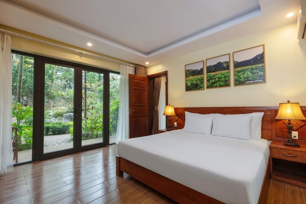 Quarto de hotel com uma cama grande, duas mesas de cabeceira com abajures, uma porta de vidro deslizante para um jardim e três quadros na parede.