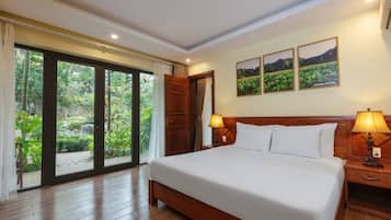 Una habitación de hotel con una cama grande, dos mesitas de noche con lámparas, una puerta corrediza de vidrio a un jardín y tres cuadros en la pared.