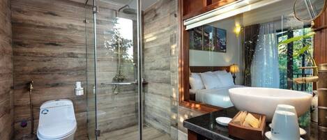 A modern bathroom with a glass-enclosed shower, a toilet, a large sink, and a bed in the adjacent room.