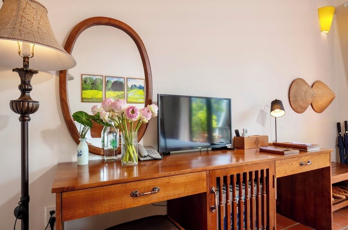 Un escritorio de madera con una televisión, una lámpara, un espejo y un florero con flores.