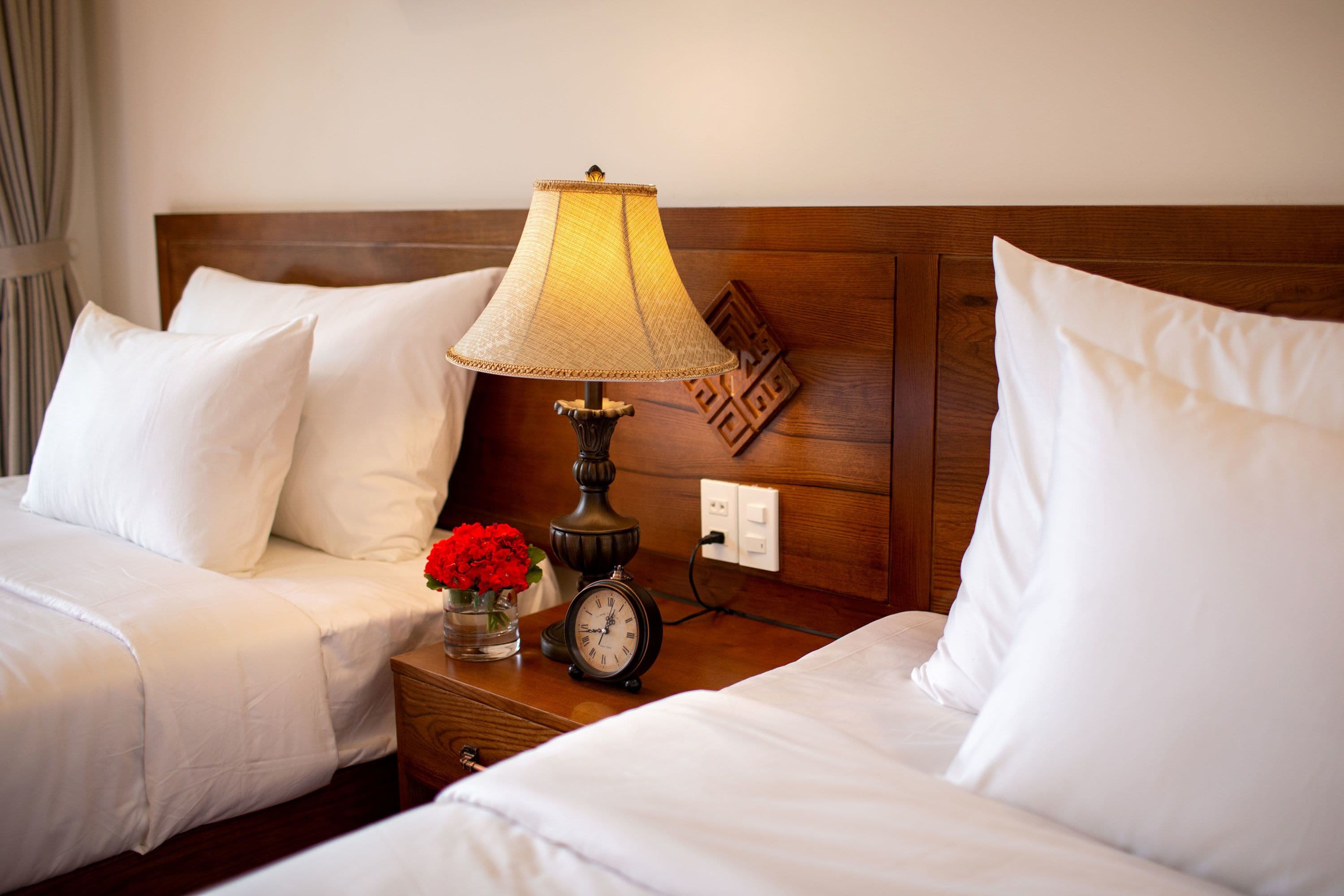 Habitación de hotel con dos camas, una mesita de noche con lámpara, un reloj y un jarrón con flores.