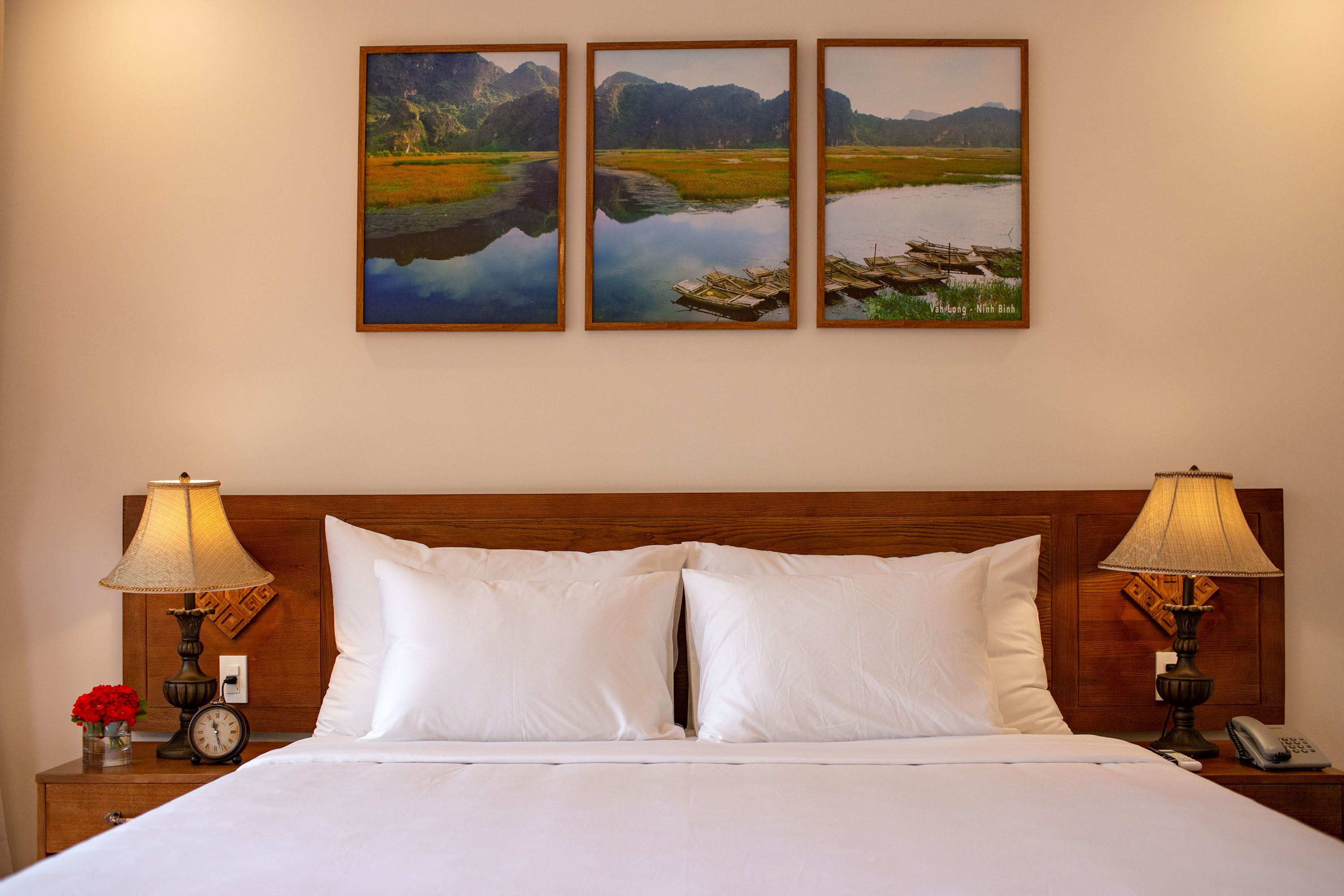 A hotel room with a wooden headboard, white bedding, two bedside lamps, a clock, and three framed landscape paintings on the wall.