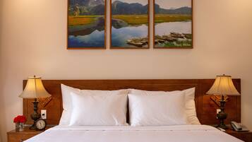 A hotel room with a wooden headboard, white bedding, two bedside lamps, a clock, and three framed landscape paintings on the wall.