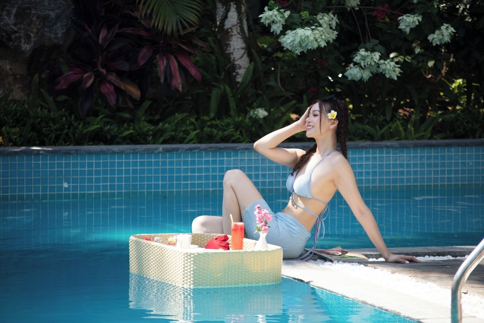 A woman sitting by a pool with a drink and a basket, surrounded by greenery.