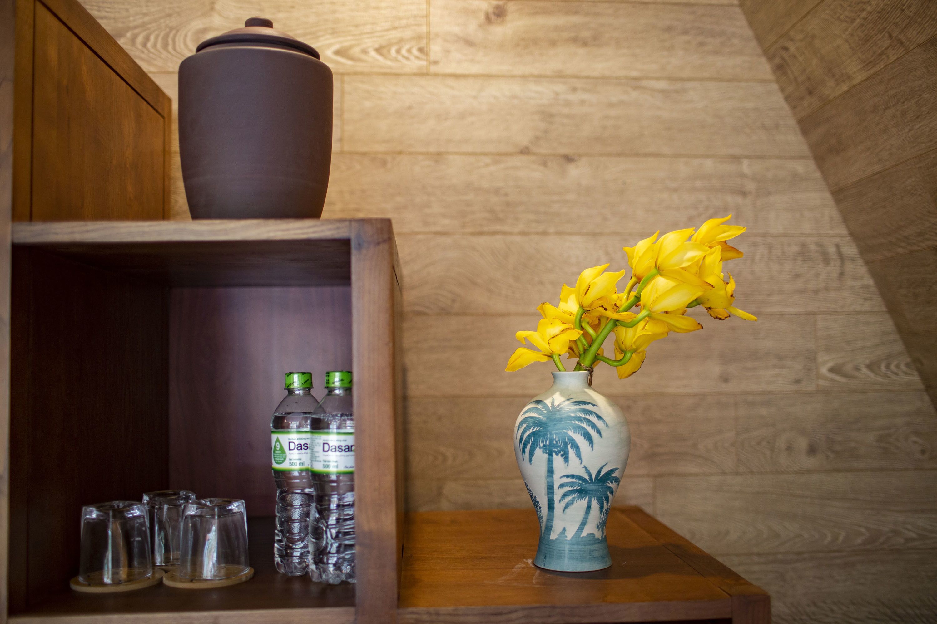 A wooden shelf with a water bottle, glasses, and a vase with yellow flowers.