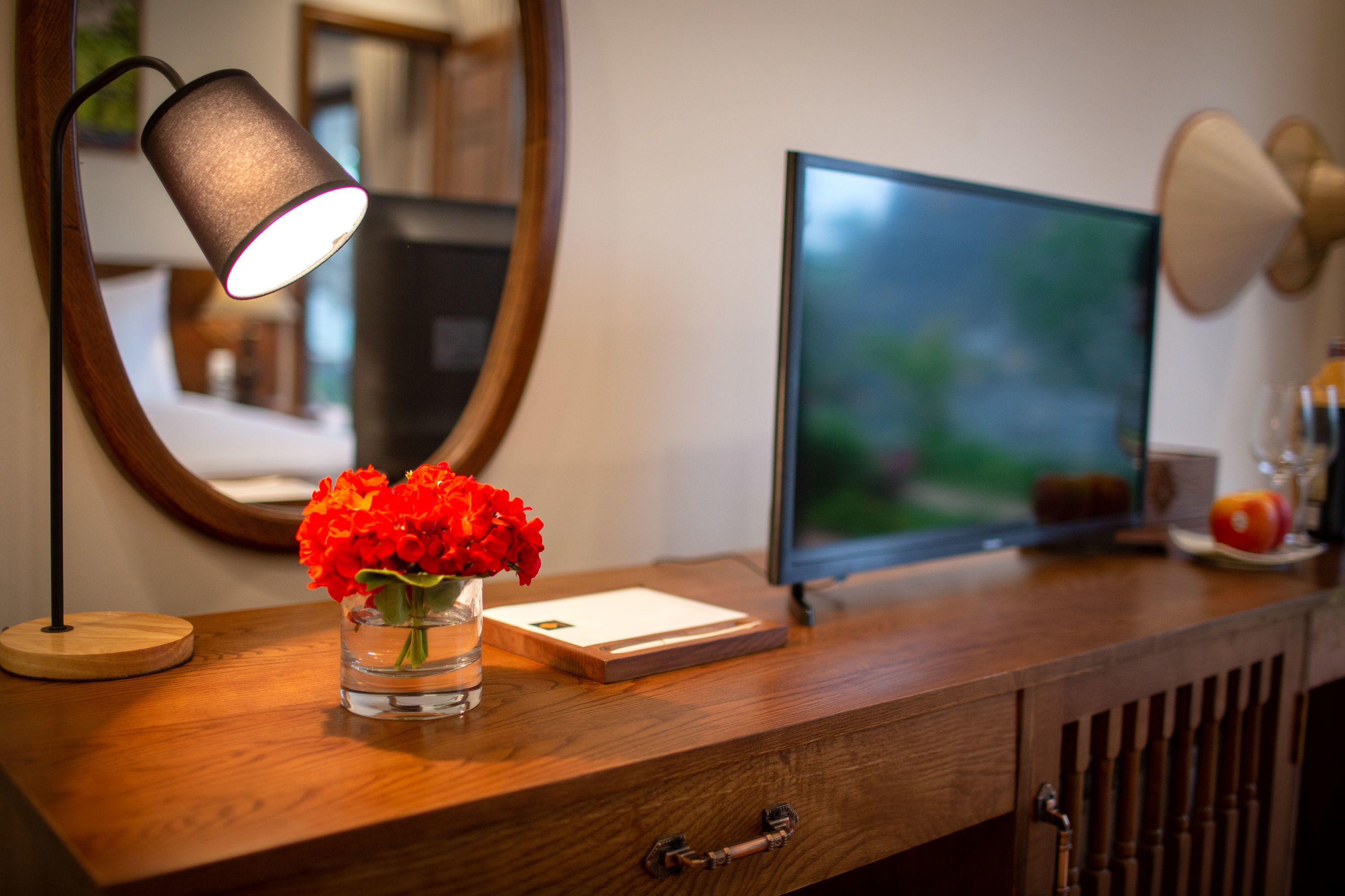 A wooden desk with a vase of red flowers, a lamp, a framed picture, and a computer monitor.