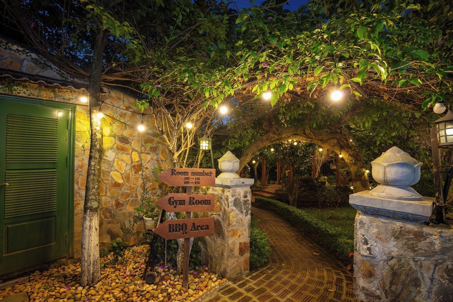 Un arco de piedra que da acceso a un patio con un cartel que indica los números de las habitaciones, un gimnasio y una zona de barbacoa.