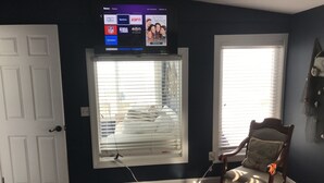 A room with a television mounted above a window, a wooden chair, and a bed with a blue blanket.