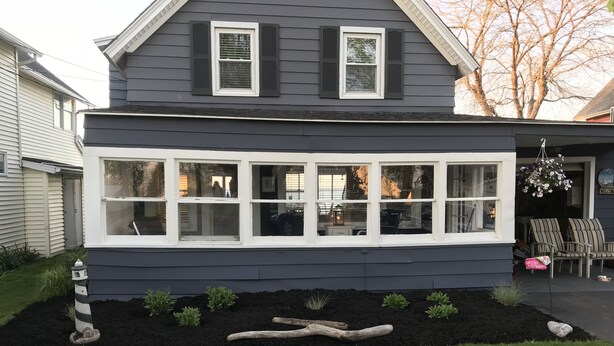 A house with a large front window, a small porch with chairs, and a garden with a wooden log and plants.