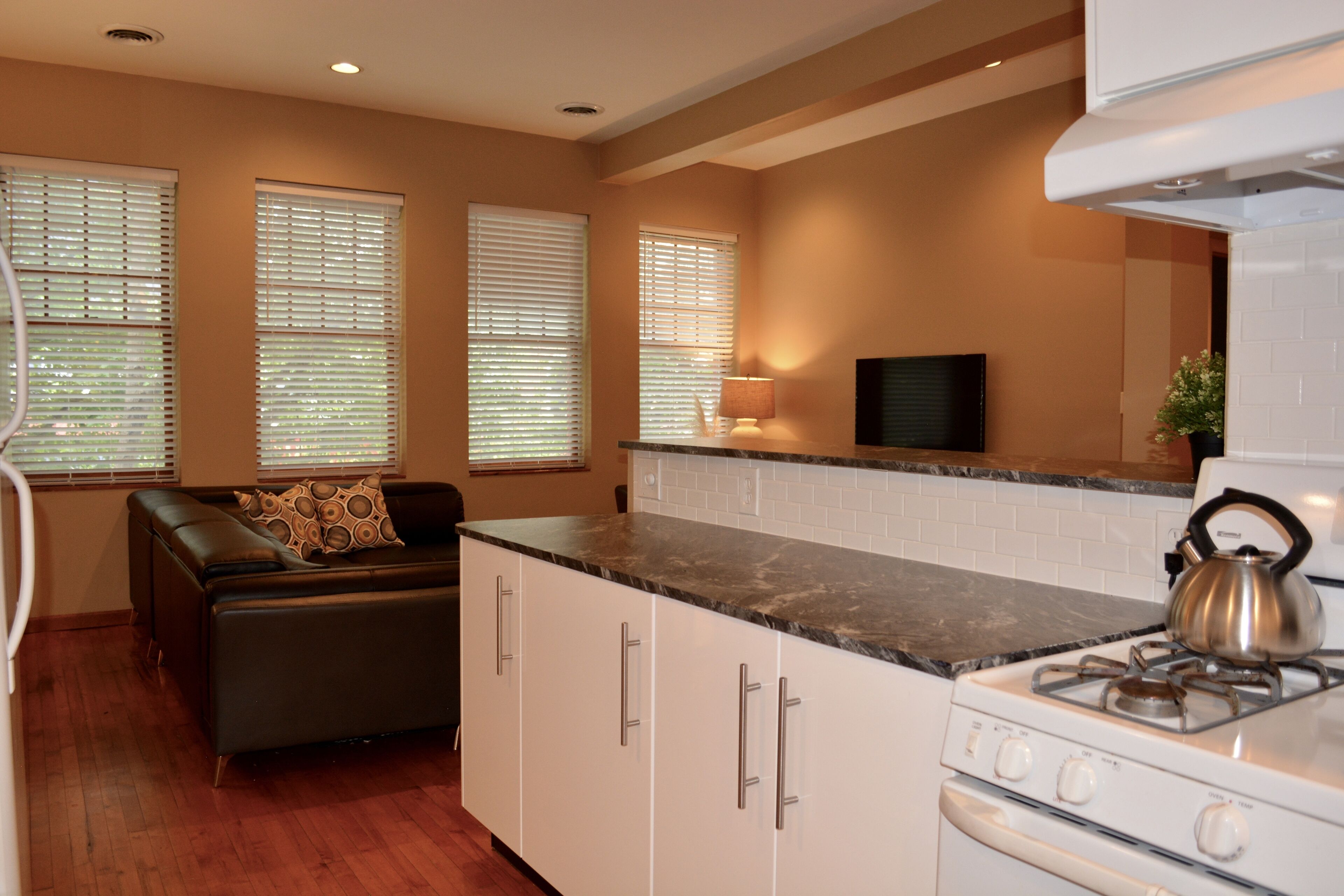 A compact kitchen with a stove, oven, and white cabinets. There are two windows with blinds, a sofa with cushions, and a small table with a lamp.