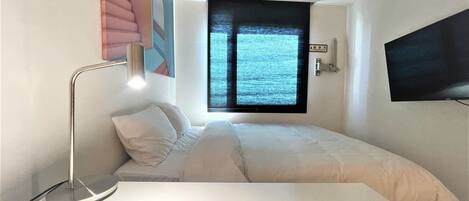 A bedroom with a bed, a desk, a chair, a desk lamp, a window with blinds, and a flat-screen TV mounted on the wall.