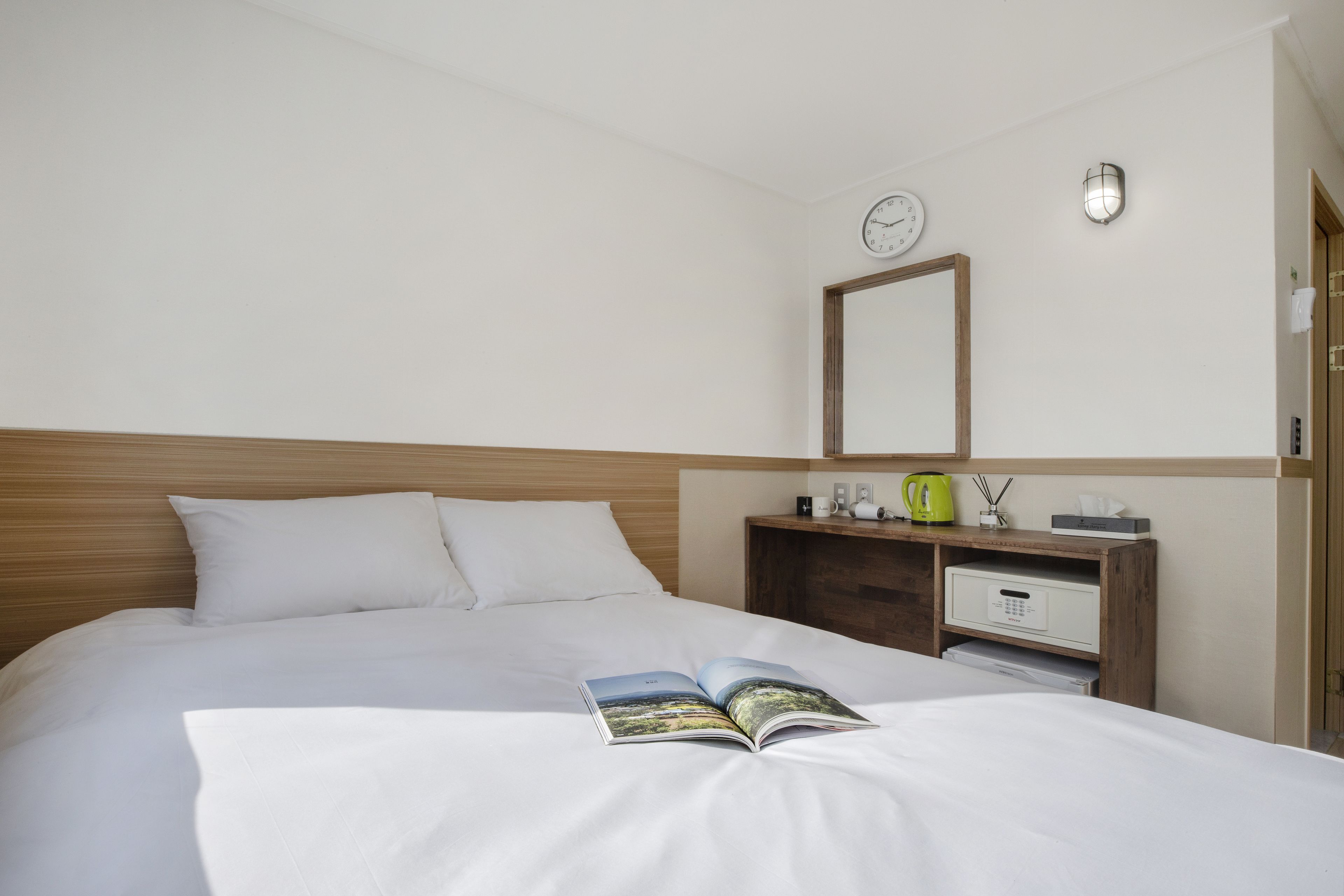 A neatly made bed with a book on it, a bedside table with a kettle, a clock, and a mirror on the wall.