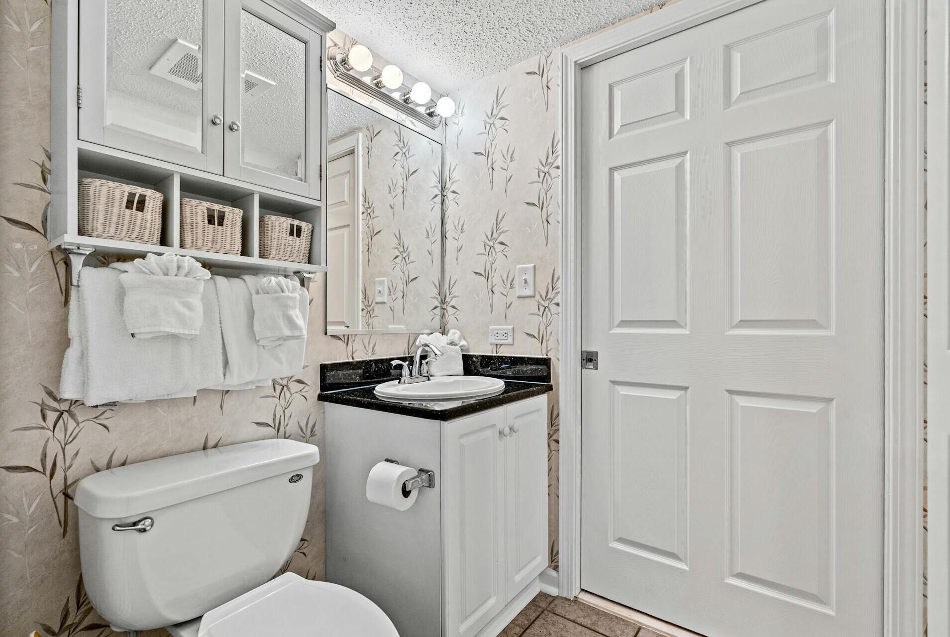 A bathroom with a toilet, sink, and mirror. There are towels hanging on the wall and a shelf above the sink.