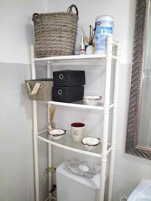 Un baño con pared de azulejos blancos, inodoro blanco, estante blanco con cajas de almacenamiento, una taza decorativa y un espejo grande.