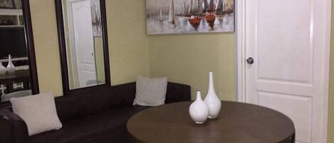 Una sala de estar con un sofá marrón oscuro, una mesa redonda de madera con tres jarrones blancos, y un cuadro con barcos a vela en la pared.