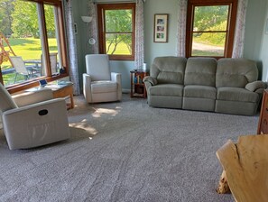 A living room with a sofa, armchair, and a coffee table. There is a window with a view of a garden.