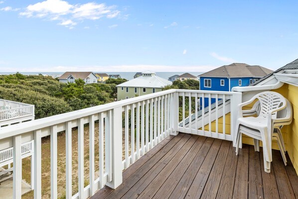 Una terraza de madera con barandal blanco, una casa azul al fondo y vista al océano.