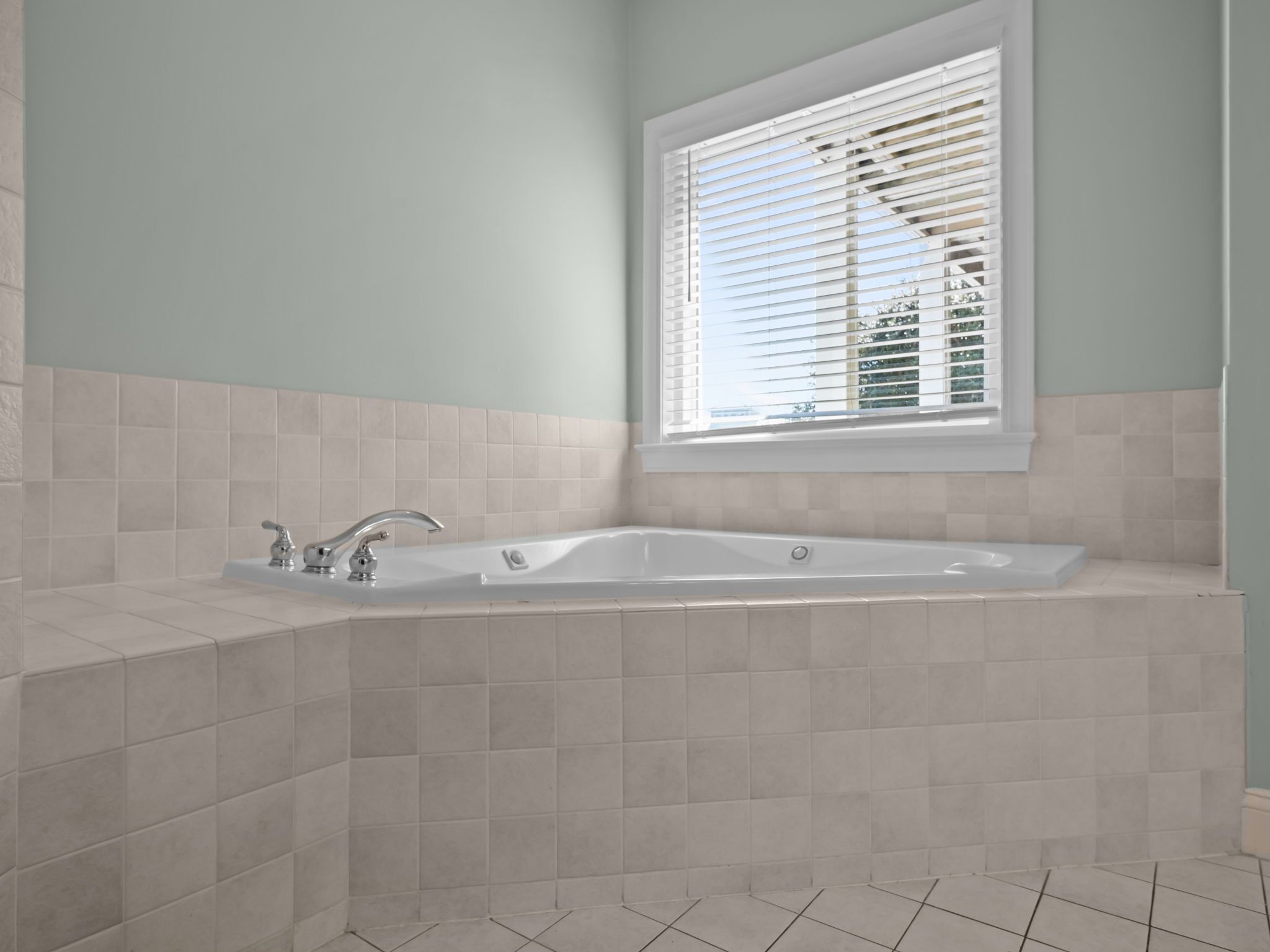A bathroom with a freestanding bathtub, a window with blinds, and tiled walls and floor.