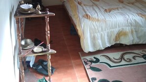 Een kleine, eenvoudige kamer met een bed, een houten tafel, een kaart aan de muur en een klein plankje met diverse spullen.