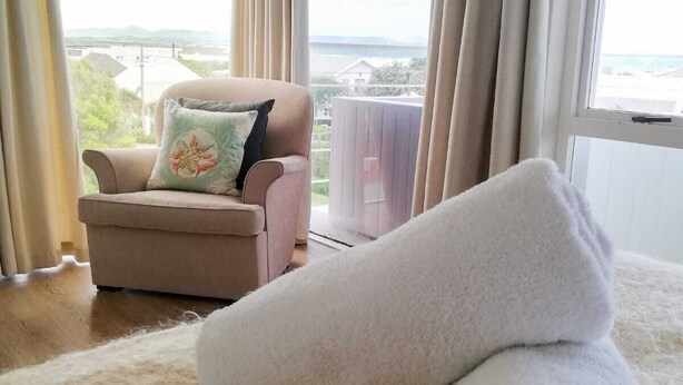 Ein Zimmer mit einem beige farbenen Sofa, einem aufgerollten weißen Handtuch und Blick auf ein Gewässer durch große Fenster.