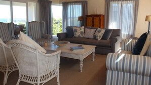Ein gemütliches Wohnzimmer mit gestreiften Sesseln und einer Couch, einem Holztisch und Blick ins Freie durch große Fenster.