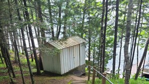 A concrete pathway leading to a small wooden shed in a wooded area near a body of water. - Bishop Lake Three Bedroom Cabin  (Crandon)