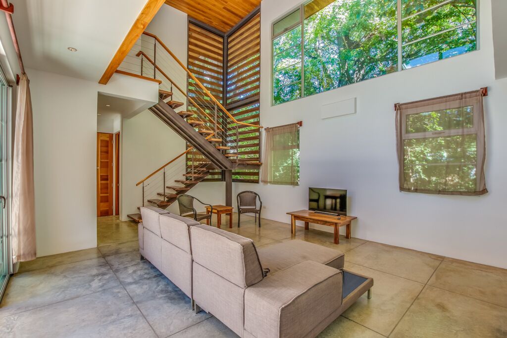 Un'ampia zona giorno con una moderna scala, un divano, una TV e grandi finestre che si affacciano sul verde.