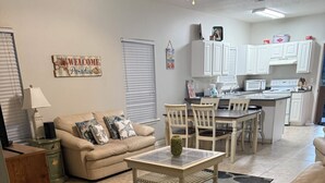 A living room with a beige sofa, a coffee table, and a dining area with chairs and a table. There is a kitchen in the background with white cabinets and a countertop.