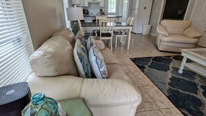 A living room with a beige sofa, a coffee table with a glass bottle, and a dining area with a white table and chairs.