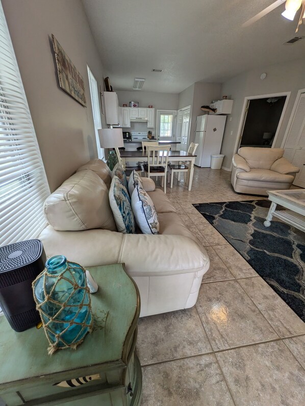 A living room with a beige sofa, a coffee table with a glass bottle, and a dining area with a white table and chairs.