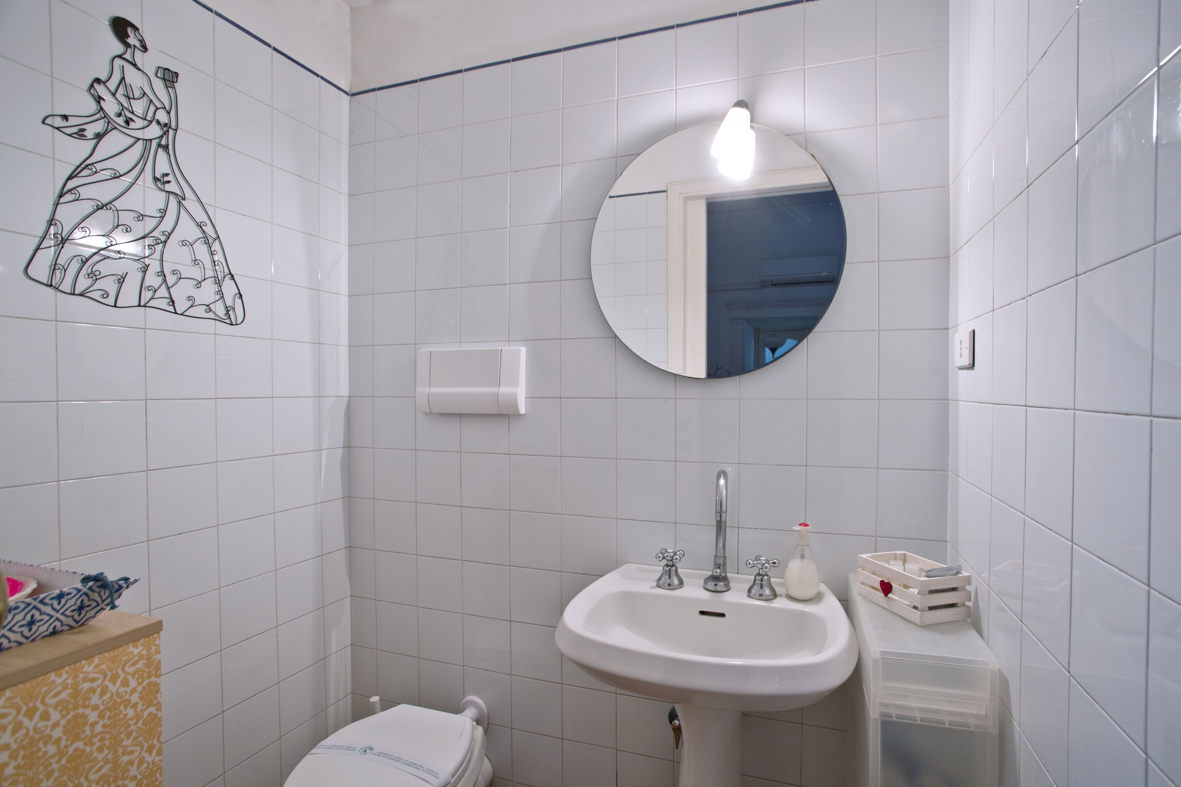 Une salle de bain avec des carreaux blancs, un miroir rond, un lavabo à deux robinets et des toilettes.