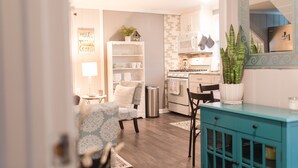 A modern kitchen with white cabinets, a stainless steel refrigerator, and a microwave. There is a dining area with a blue patterned chair and a teal cabinet. - North End Beach Cottage Apartment - One block from beach (Virginia Beach)