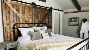 Une chambre avec une tête de lit en bois, une couvre-lit blanche et un plaid moelleux. On y trouve une table de chevet avec un tiroir et une petite lampe de table. Un tableau encadré est accroché au mur, et un peignoir blanc est suspendu à un crochet.