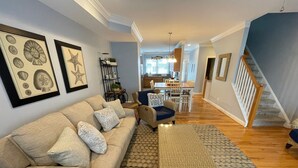 Un salon spacieux avec un canapé beige, des coussins à motifs, un pouf en osier et, au fond, un coin repas.