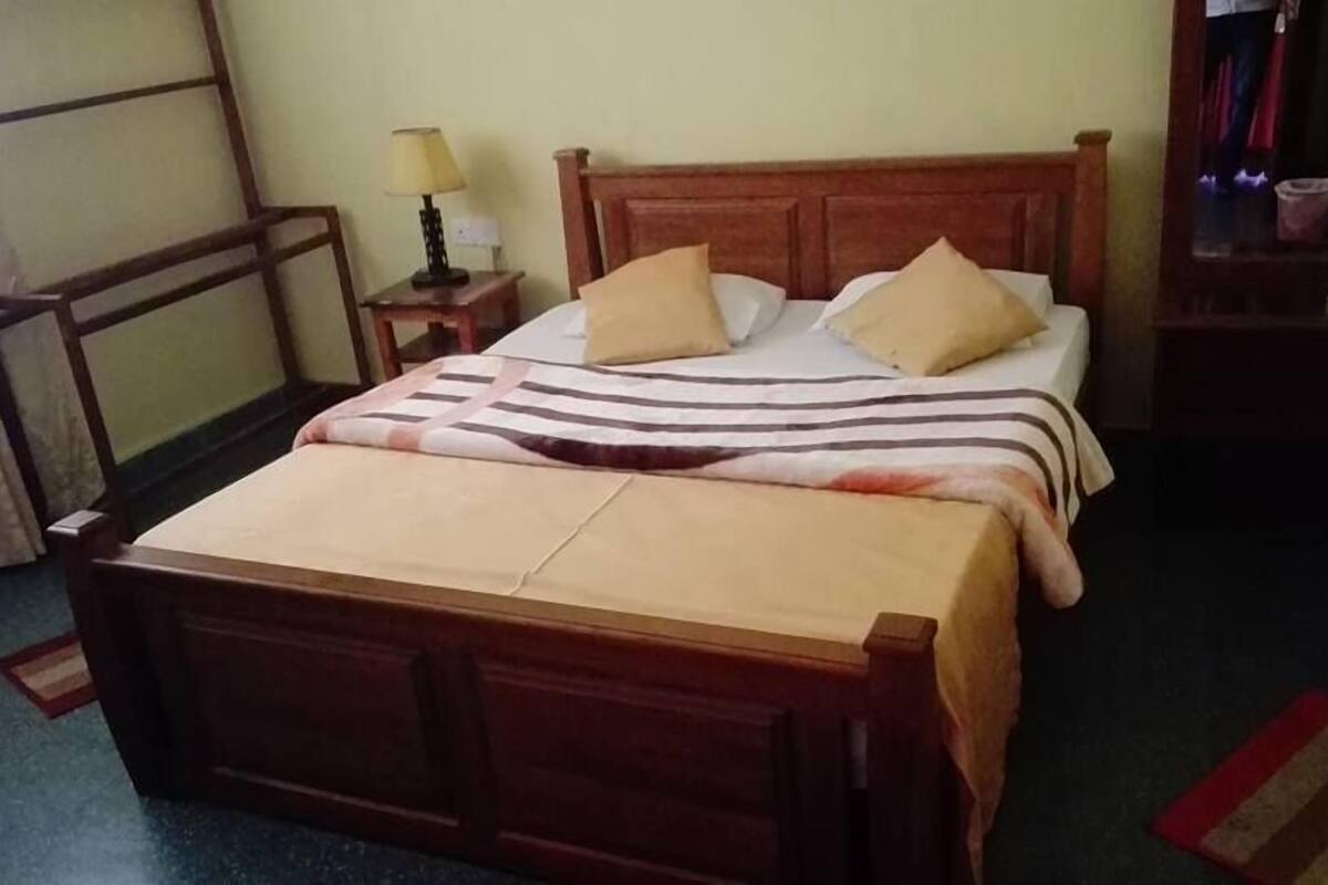 Una cama individual con estructura de madera, una colcha a rayas y dos almohadas. A la izquierda de la cama, una mesita de noche con una lámpara.