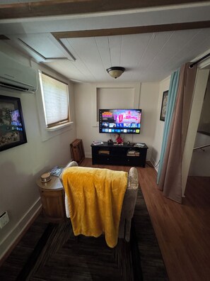 Ein Wohnzimmer mit Fernseher, einem kleinen Tisch, einer Couch und einem Fenster mit Jalousien.