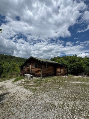 A wooden cabin with a gravel driveway, surrounded by trees and under a partly cloudy sky.