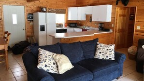 A cozy cabin interior with a wooden ceiling, a blue sofa with patterned cushions, a white kitchen area with a microwave, and a bear decoration. - Cute Lakefront Cottage on Lake Michigan.   (Mears)